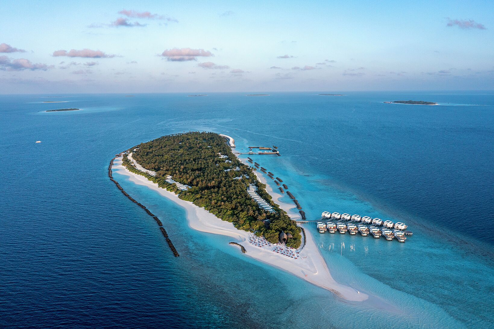 An aerial view of a tropical island with a long stretch of white sand beach, a dense forested area, and a row of overwater bungalows.