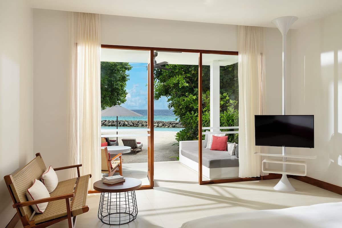 A modern hotel room with a large sliding glass door leading to a beach view, a flat-screen TV, a wicker chair, and a small round table.