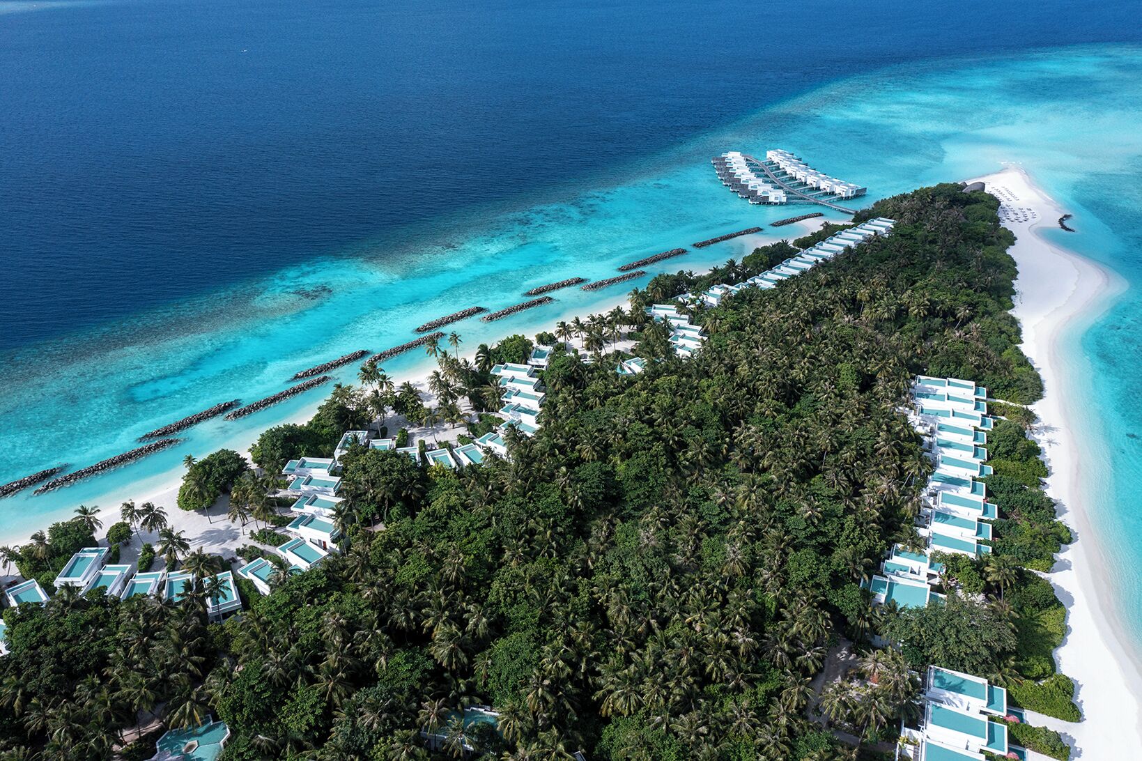 Vista aérea de un complejo turístico con múltiples bungalows sobre el agua, una playa y aguas turquesas cristalinas.