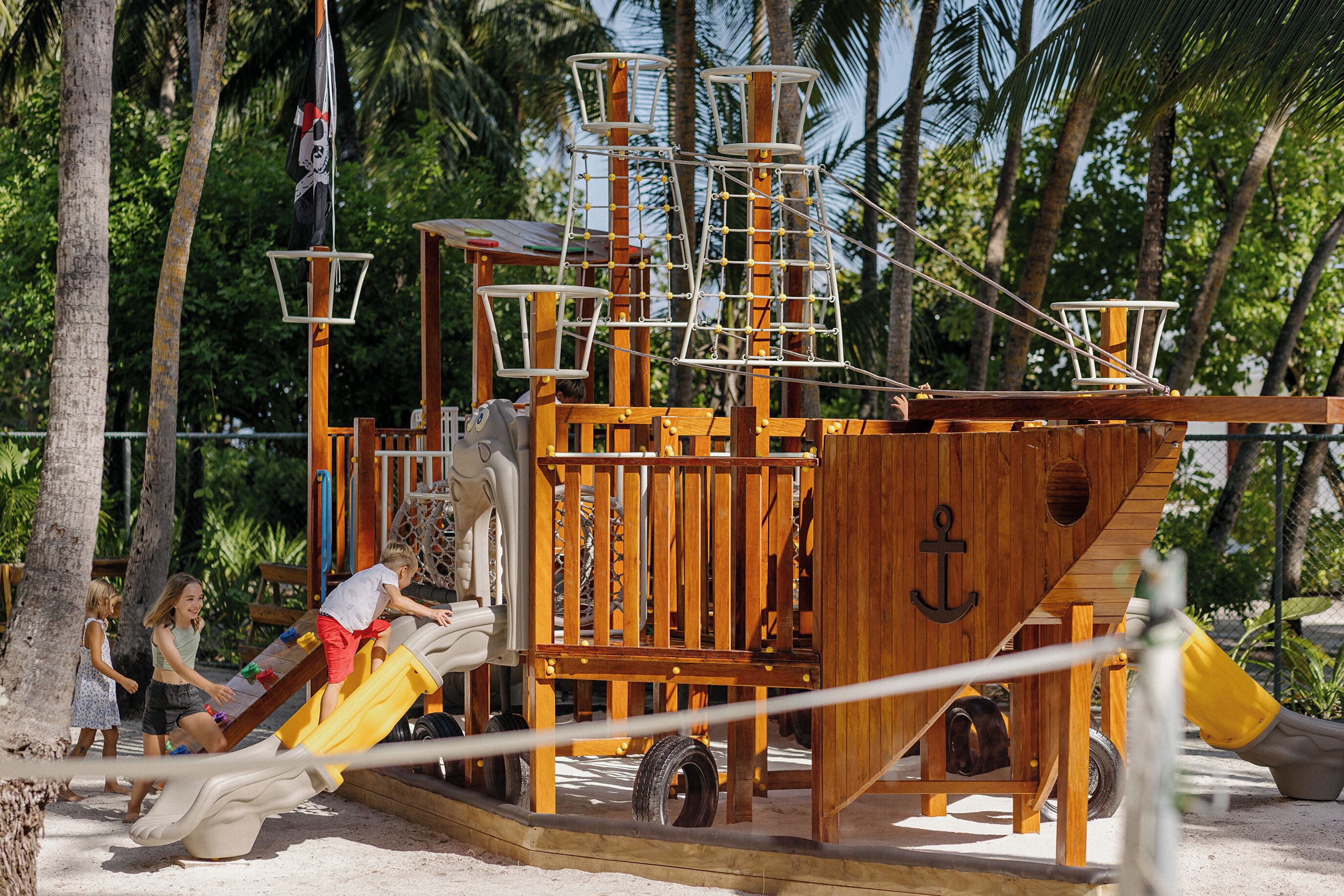 A wooden pirate ship play structure with slides and climbing nets, surrounded by palm trees.