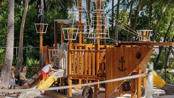 A wooden pirate ship play structure with slides and climbing nets, surrounded by palm trees.
