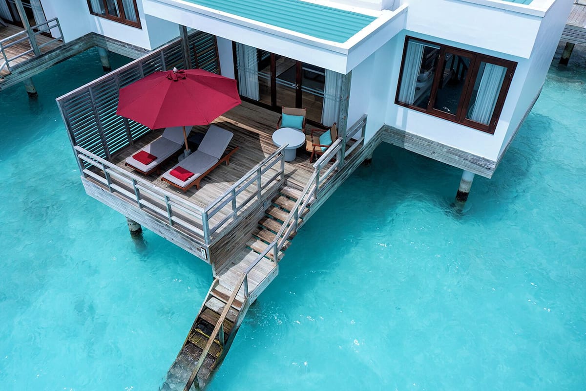 An overwater bungalow with a wooden deck, lounge chairs, and a red umbrella.