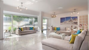 A spacious living room with a large window, a sofa set, a coffee table, and a chandelier. - Milos Hotel (Milos)