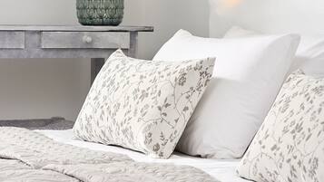 A bedroom with a bed, floral-patterned pillows, a nightstand with a vase of orchids, and a white wall.