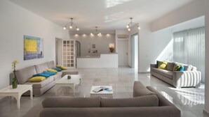 A modern living room with a sofa, coffee table, and a dining area. - Milos Hotel (Milos)