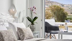 A bedroom with a bed, pillows, a nightstand, a potted orchid, a chair, and a table with a bottle and glass. - Milos Hotel (Milos)