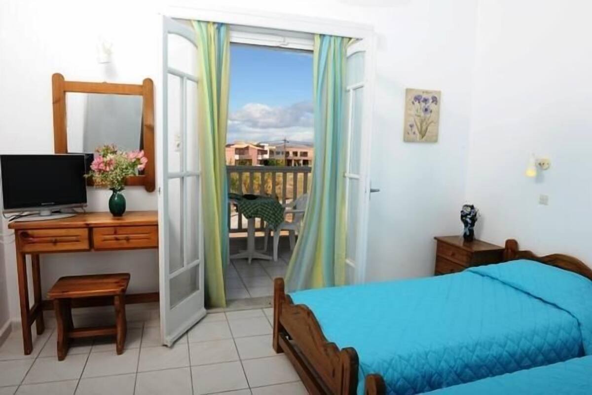 A bedroom with a bed, a desk with a TV, a chair, a small table, and a balcony with a view of buildings.