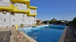 A swimming pool with a stone deck, a yellow and white building with balconies, and a clear blue sky. - Kamaria Apartments (Hersonissos)