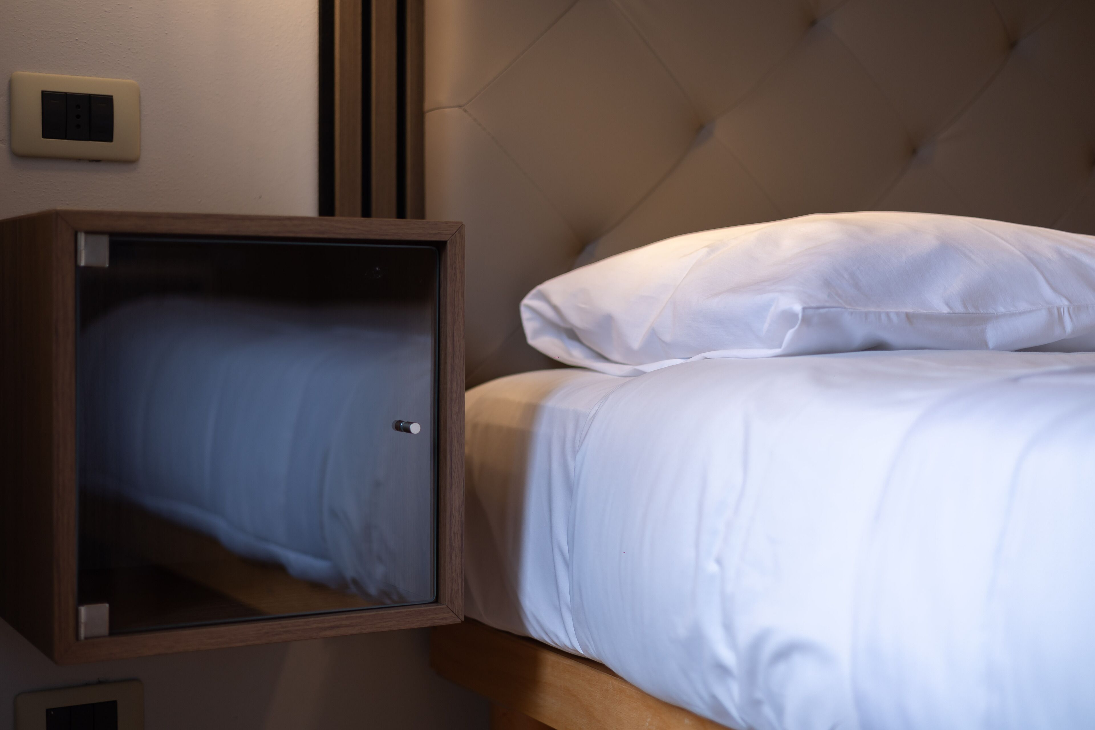 A bedroom with a bed, a wooden nightstand, and a wall-mounted light switch.