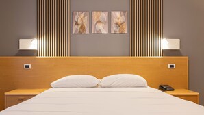 A hotel room with a wooden headboard, a large bed with white bedding, two bedside tables with lamps, and three framed artworks on the wall. - La Fertè Hotel (Asti)