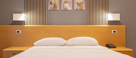 Una habitación de hotel con cabecera de madera, una cama grande con ropa de cama blanca, dos mesitas de noche con lámparas y tres obras de arte enmarcadas en la pared.