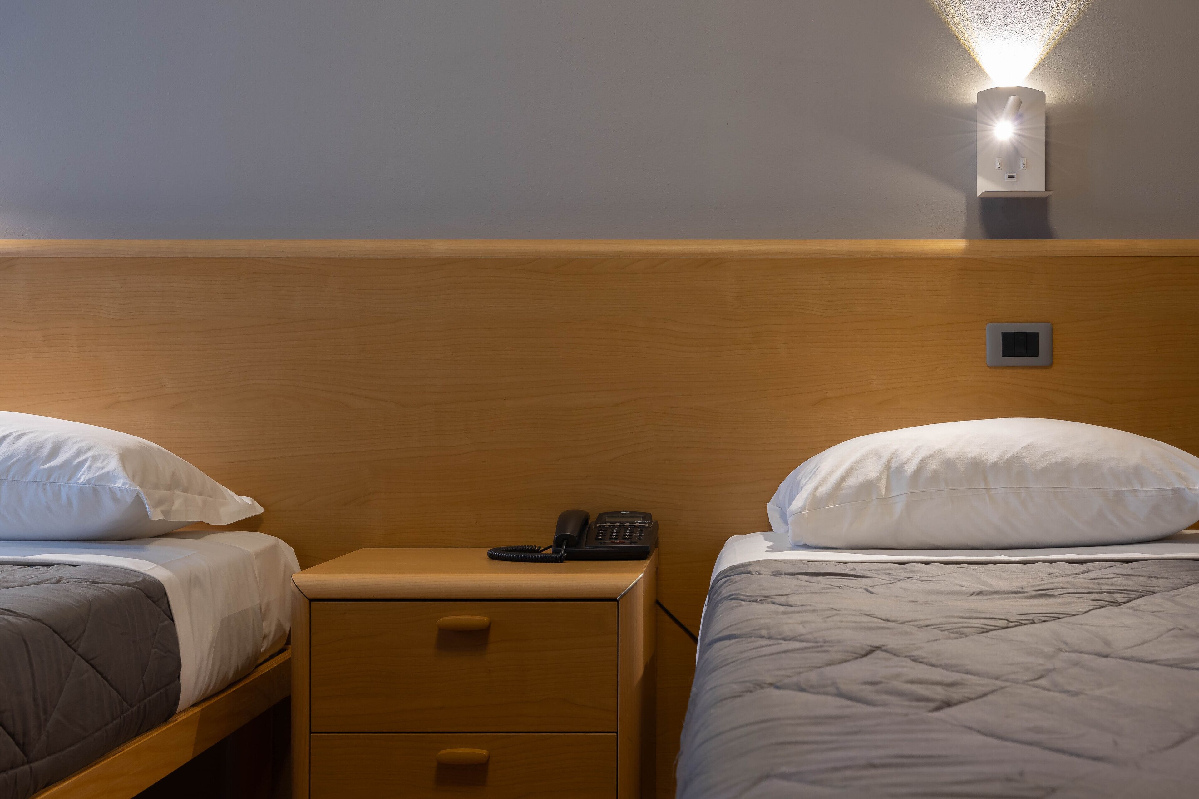 A hotel room with two beds, a bedside table with a phone, and a wall-mounted light fixture.