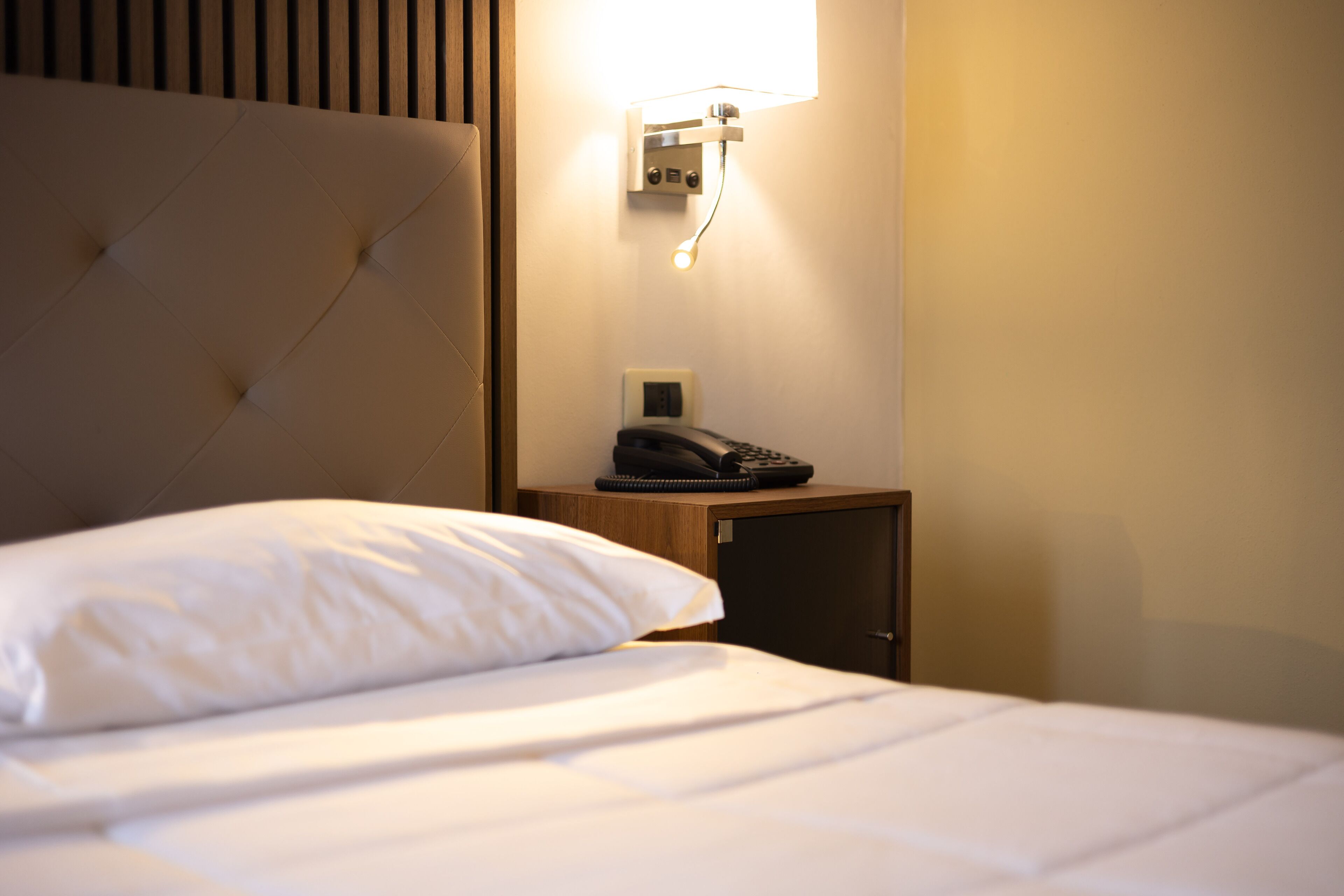 A neatly made bed with a white pillow and a wooden headboard. A bedside table with a telephone and a wall-mounted light fixture is visible.