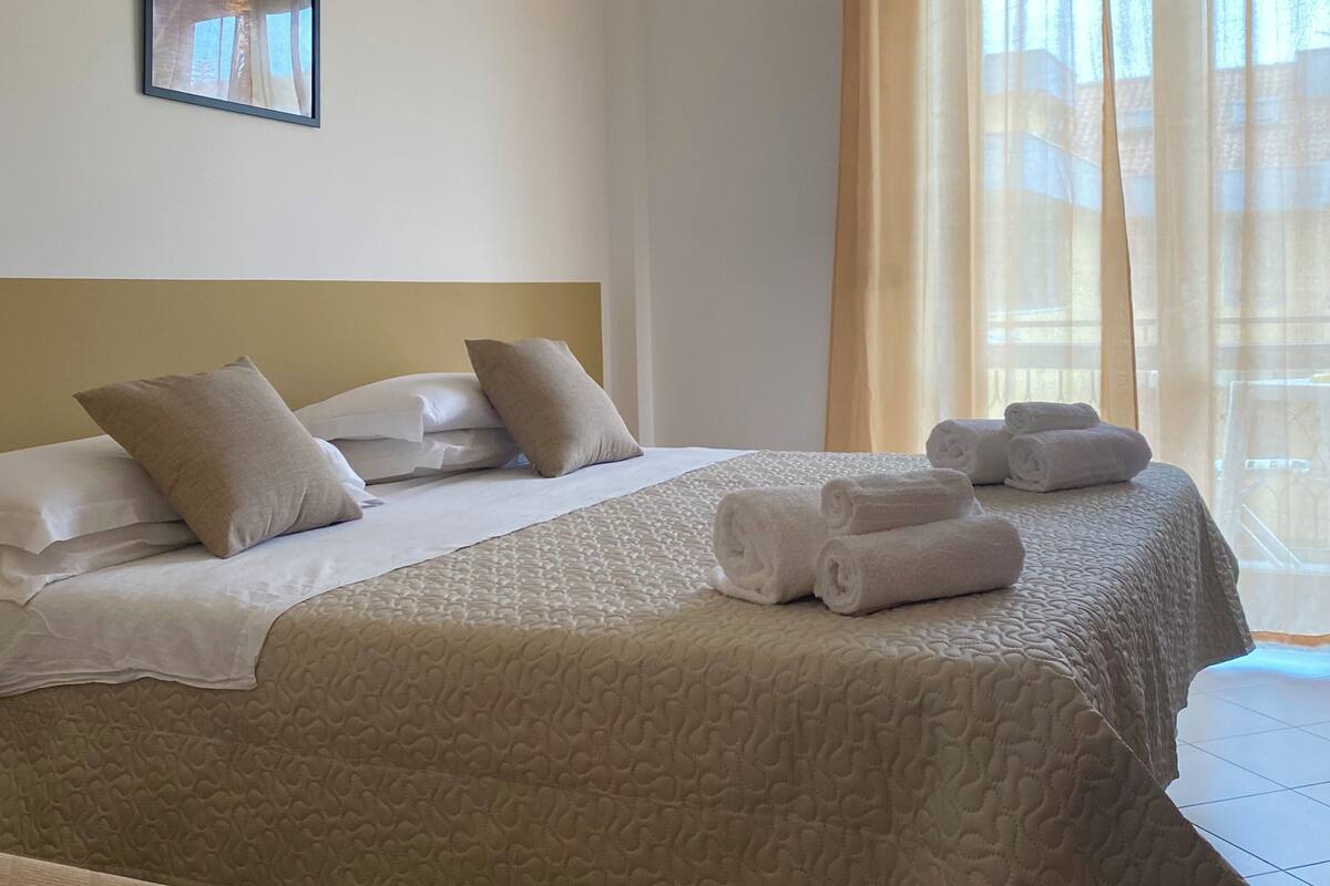 Ein Hotelzimmer mit zwei Betten, beiger Bettwäsche, einem Kopfteil, einem gerahmten Bild an der Wand und einem Fenster mit Vorhängen.
