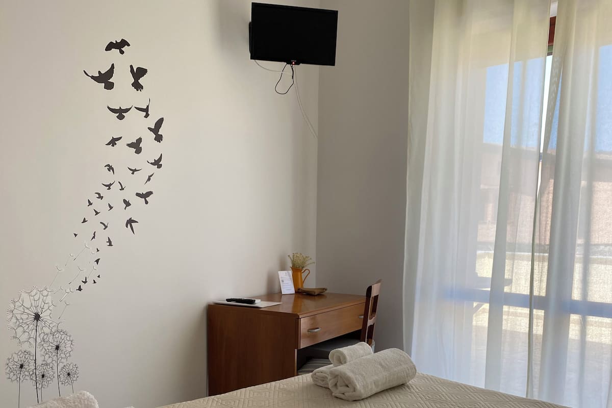 Ein Hotelzimmer mit Bett, einem Holzkleiderschrank, einem flachbildfernseher an der Wand und einem Fenster mit durchsichtigen Vorhängen.