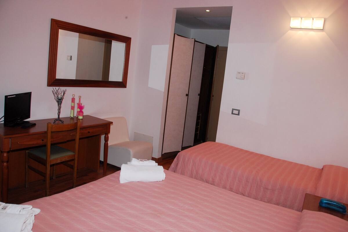 A hotel room with a bed, a desk with a chair, a mirror, and a wardrobe.