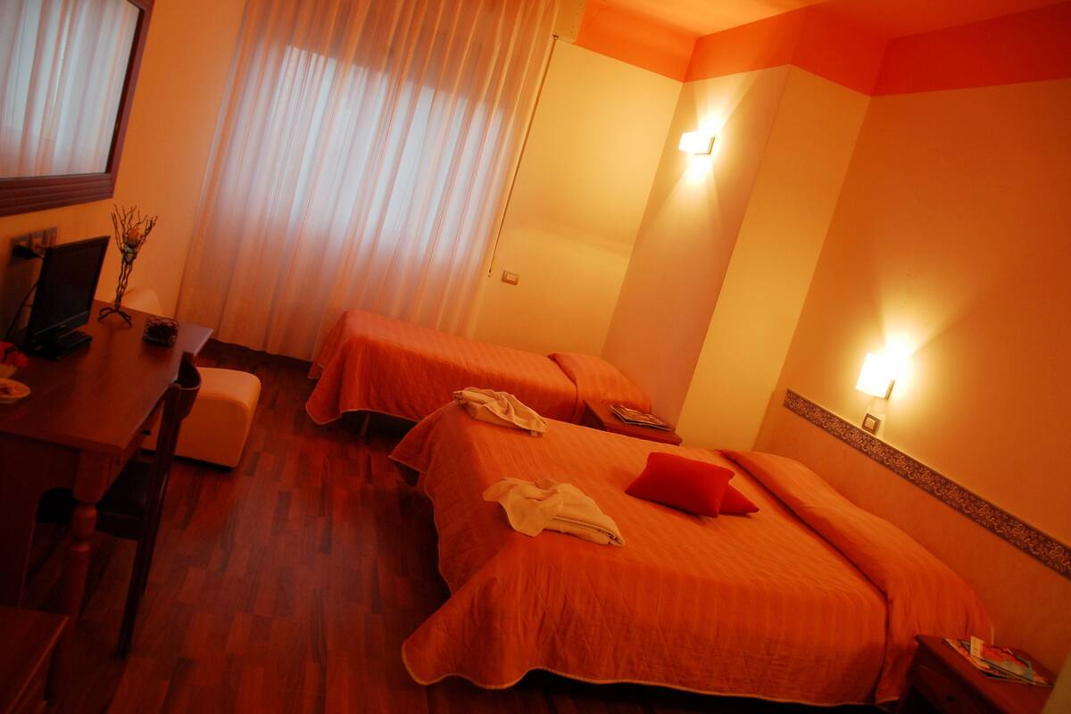 A hotel room with two beds, a desk with a chair, a small table, and a window with curtains.