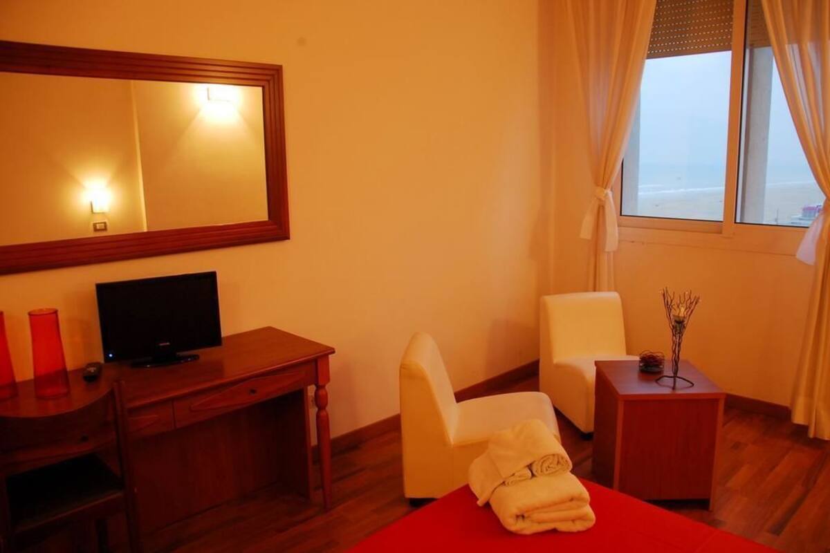 A hotel room with a wooden desk, a television, a chair, a bed with a red bedspread, and a window with curtains.