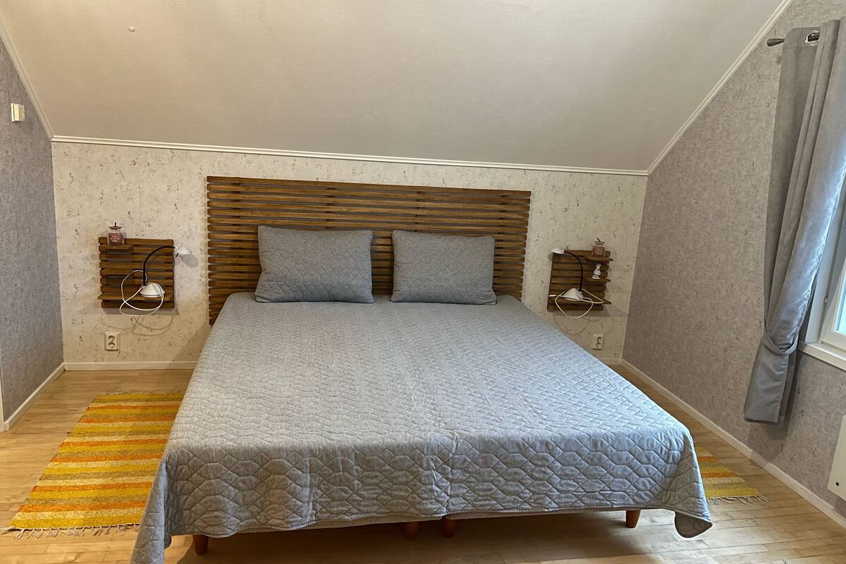 Un dormitorio con una cama grande, cabecera de madera, dos mesitas de noche con lámparas, una alfombra a rayas y una ventana con cortinas.