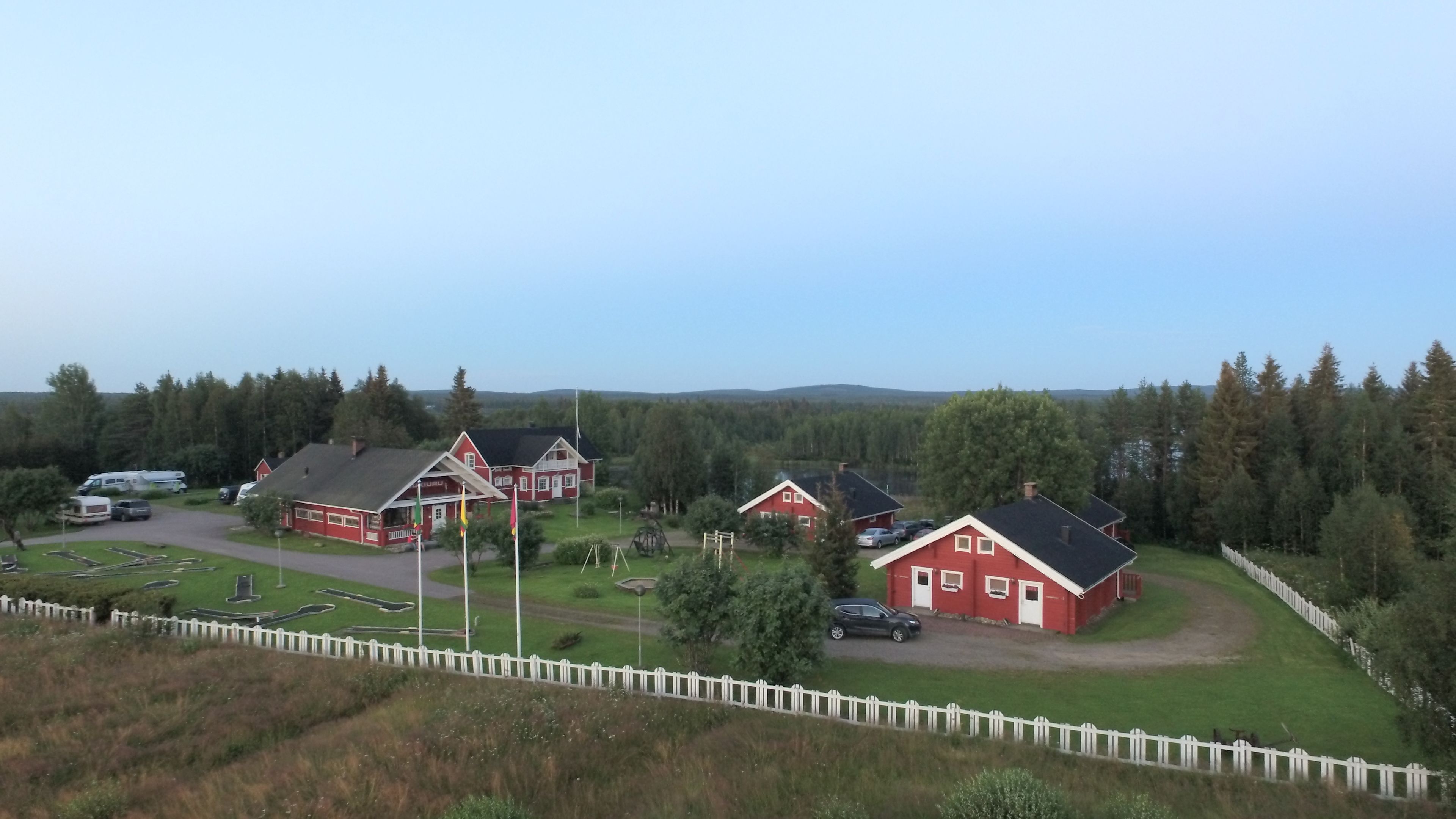 A red building with a white fence in front, surrounded by greenery and a clear sky.