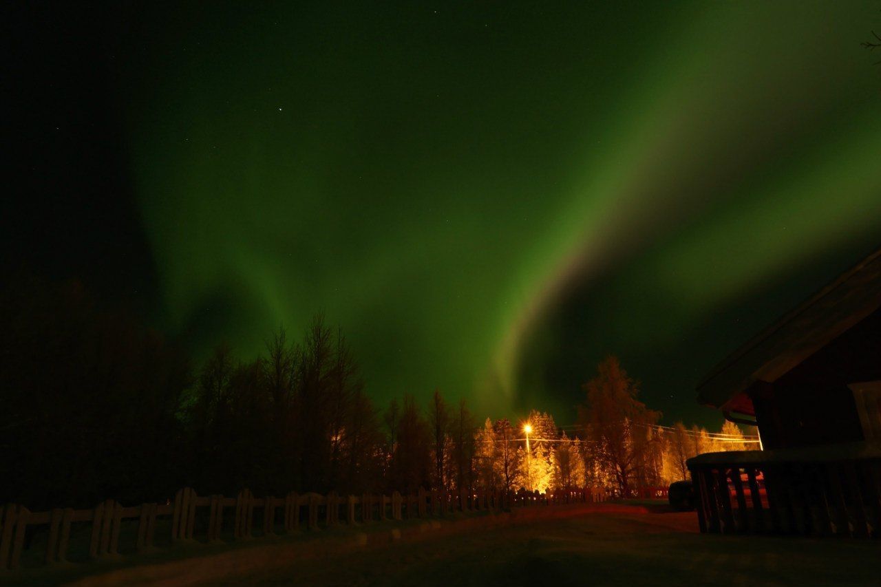 A night scene with the aurora borealis over a rural area, featuring a wooden fence, a lit building, and a road.