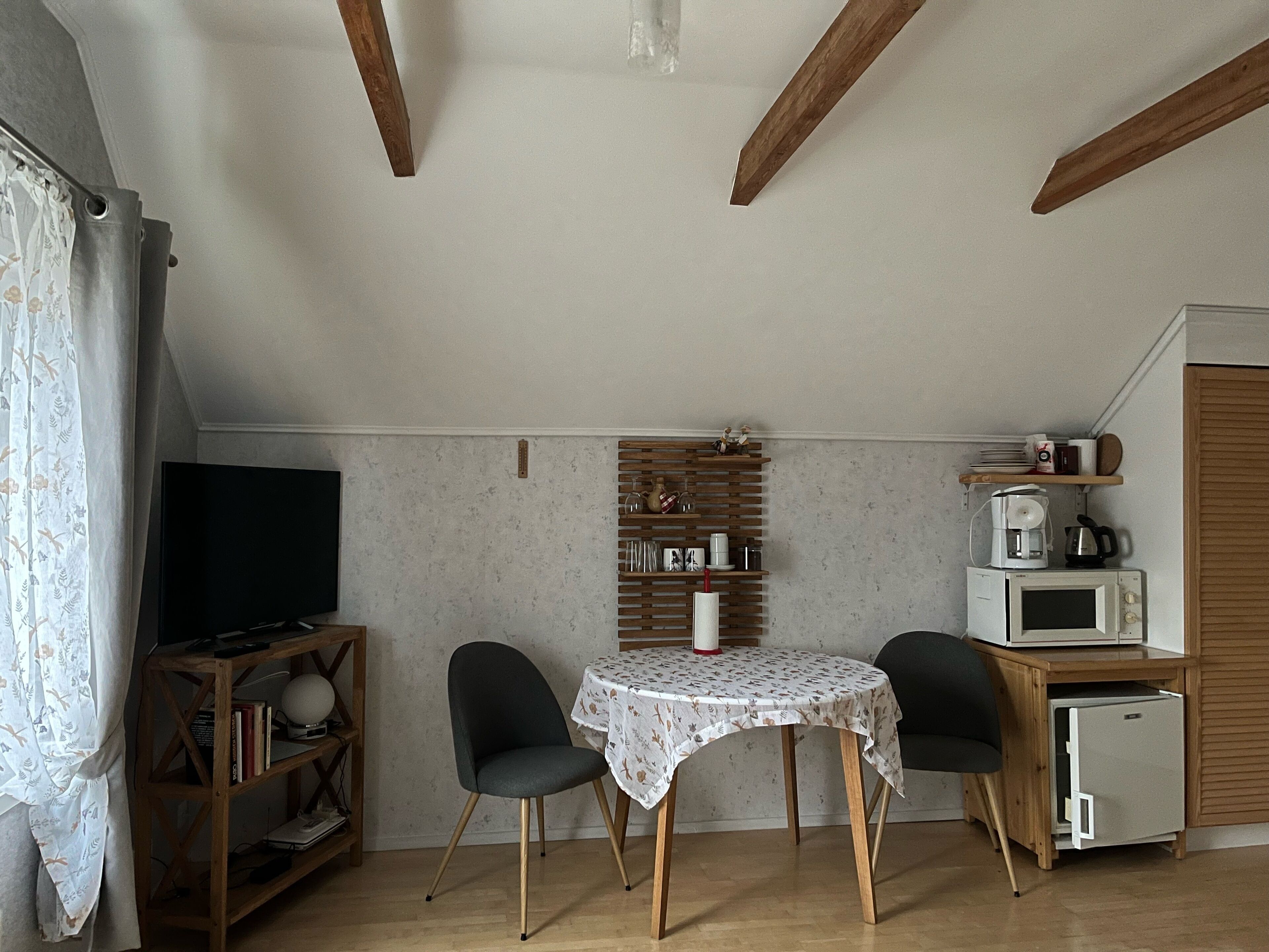 A cozy room with a wooden ceiling, a small table with chairs, a television, a bookshelf, and a kitchen area with a microwave and coffee maker.