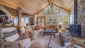 A cozy living room with a stone fireplace, wooden ceiling beams, and large windows offering natural light.