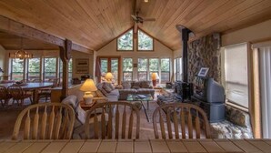 A cozy cabin interior with a wooden ceiling, stone fireplace, and a dining area with wooden furniture.