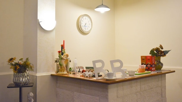 A hotel reception area with a clock, a counter with various items, a vase with flowers, and a pendant light.