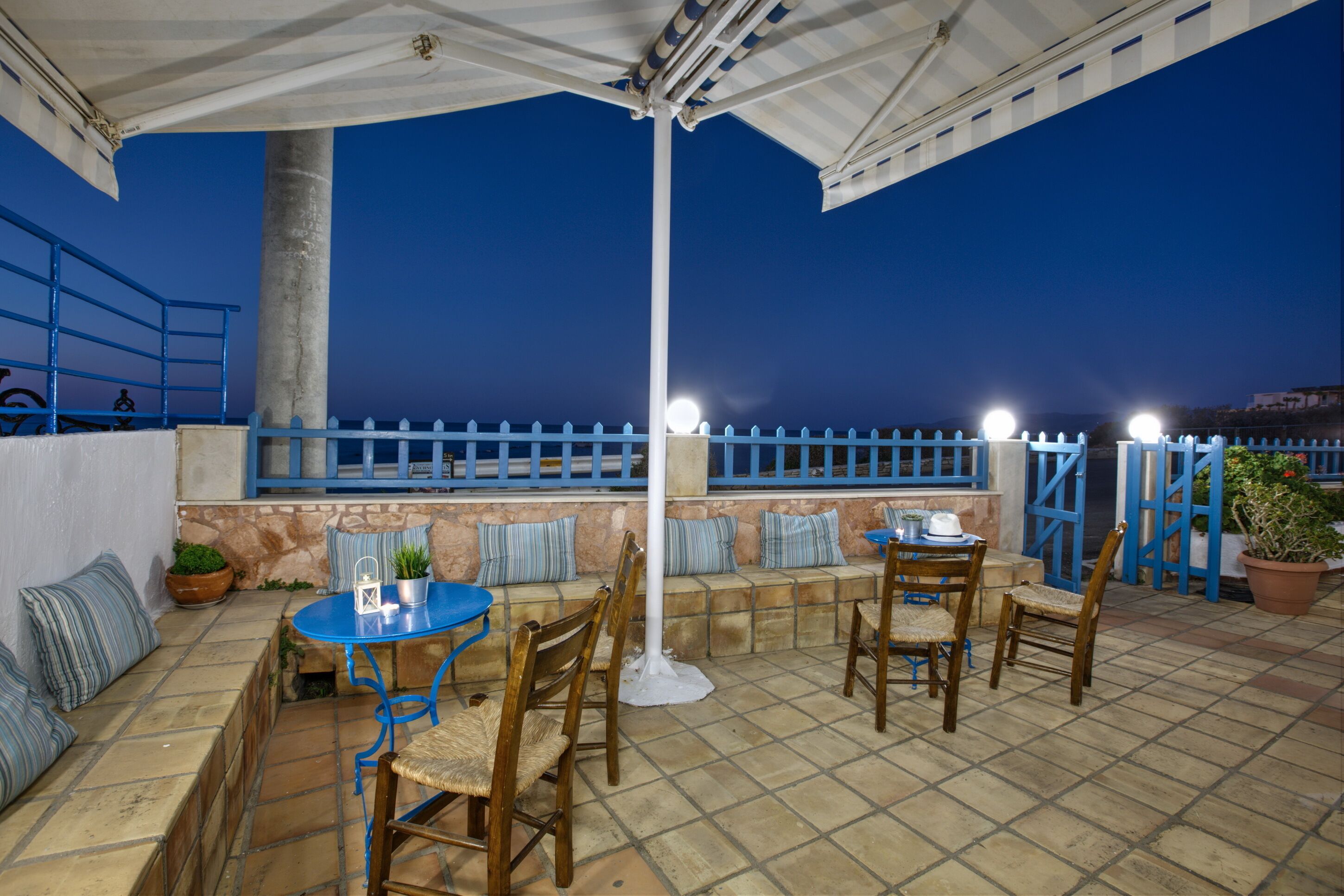 An outdoor patio area with a blue table and chairs, a white pole, and a view of the sea at dusk.
