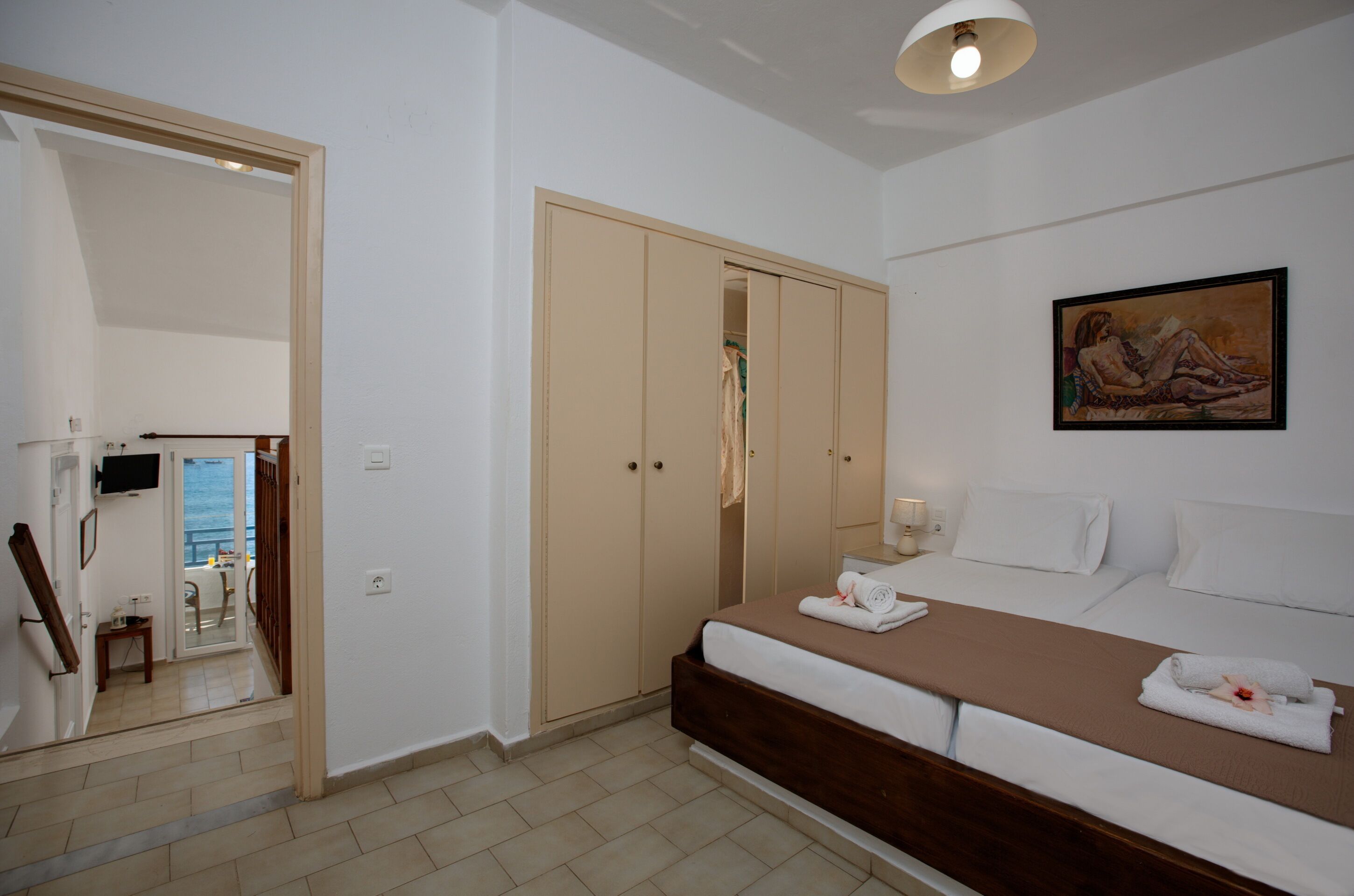 A hotel room with a bed, a wardrobe, a painting on the wall, and a view of the sea through an open door.