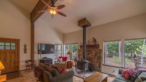 A cozy living room with a stone fireplace, a ceiling fan, and a large window overlooking a forest. - Kelly 3 Bedroom Holiday Home By Tahoe Truckee (Truckee)