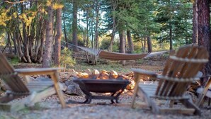 Eine Hängematze zwischen zwei Holzsesseln inmitten eines Waldes, mit einem Feuerkorb.
