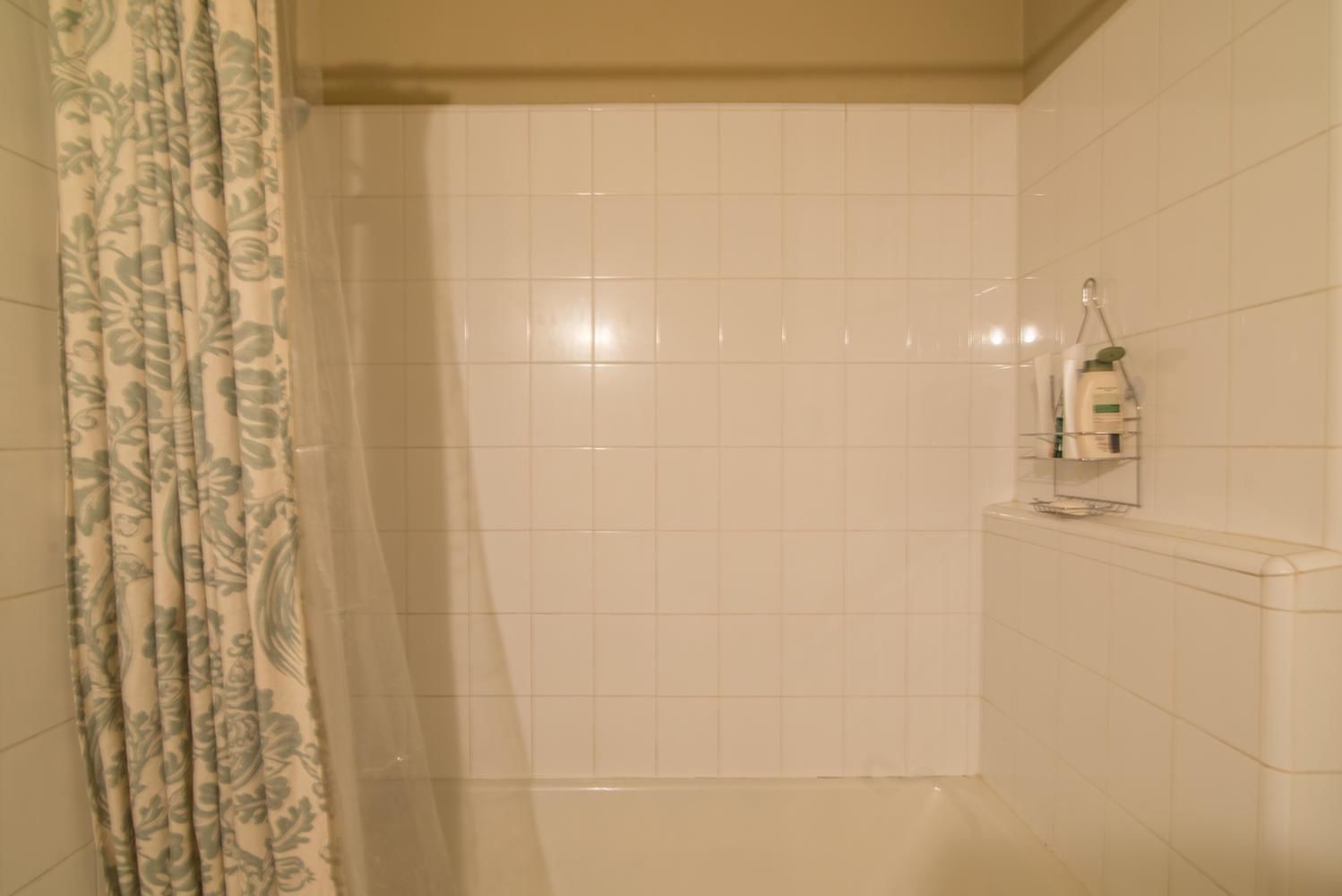 A bathroom with white tiled walls, a shower area, and a toilet with a shelf holding toiletries.