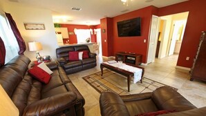 A living room with a brown leather sofa, a black leather sofa, and a wooden coffee table. There is a kitchen in the background with white cabinets and a dining area.