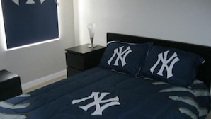 A bedroom with a bed featuring a dark blue bedspread with a white logo, two matching pillows, a small bedside table with a lamp, and a window with blinds. - Gorgeous Home with Golf Course View (Haines City)
