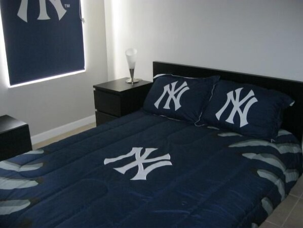 Un dormitorio con una cama que tiene una colcha azul oscuro con un logotipo blanco, dos almohadas a juego, una mesita de noche con una lámpara y una ventana con persianas.