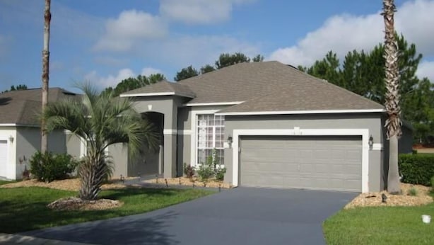 A single-story house with a garage, a driveway, and a lawn with palm trees. - Gorgeous Home with Golf Course View (Haines City)