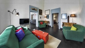 A modern living room with green sofas, a red ottoman, and a television. There are two lamps, a wooden side table, and a framed picture on the wall. - Palazzo Melfi Suite - Hotel (Comiso)