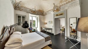 A bedroom with a large bed, a desk, and a chair. There is a window with curtains, a mirror, and a ceiling with intricate designs. - Palazzo Melfi Suite - Hotel (Comiso)