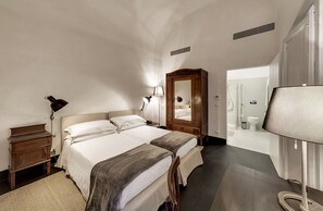 A bedroom with two beds, a wooden wardrobe, a mirror, and a bathroom visible through an open door. - Palazzo Melfi Suite - Hotel (Comiso)