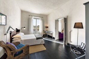 A modern hotel room with a large bed, a desk, and a chair. There is a wicker chair with a blue cushion and a small table with a lamp. - Palazzo Melfi Suite - Hotel (Comiso)