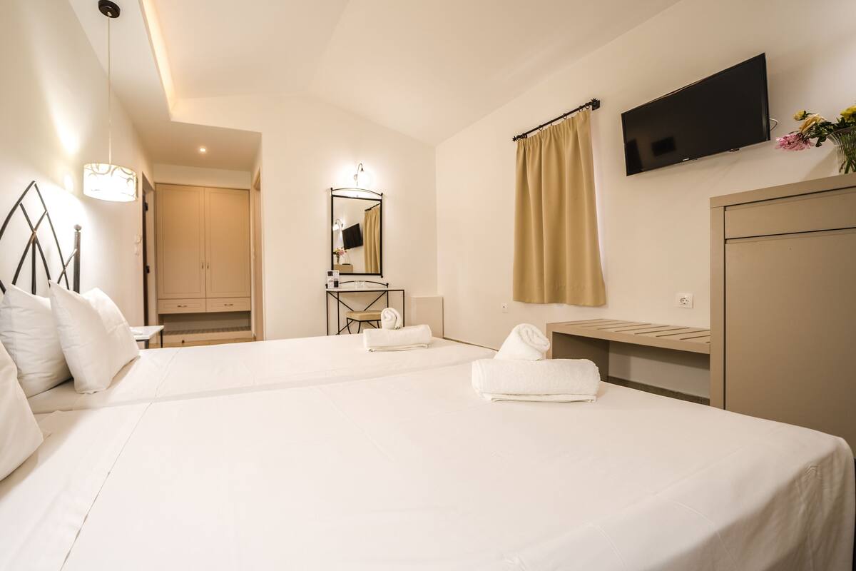 A hotel room with two beds, a desk, a mirror, a TV, and a bathroom visible in the background.