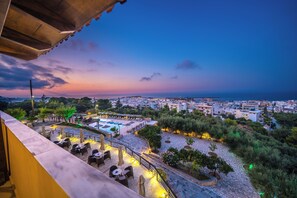 Una terraza con mesas al aire libre, piscina y vistas a la ciudad al atardecer.