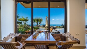 Un tablero de ajedrez sobre una mesa de madera con sillas ornamentadas, con vista a un patio con asientos al aire libre y una vista del mar.