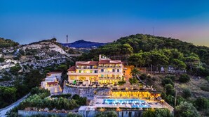 Un hotel grande de varios pisos con piscina y zona de estar al aire libre, rodeado de colinas y árboles.