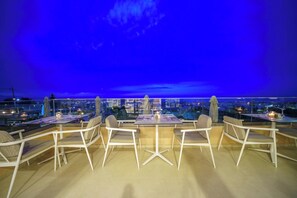 Un área de comedor en la azotea con mesas y sillas, con vista a un paisaje urbano al atardecer.