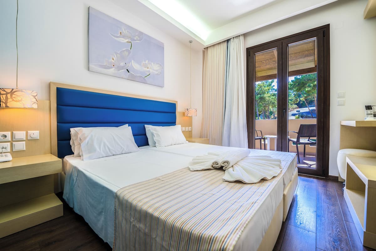 A hotel room with a large bed, a bedside table with a lamp, a painting on the wall, and a view of the outdoors through a sliding door.
