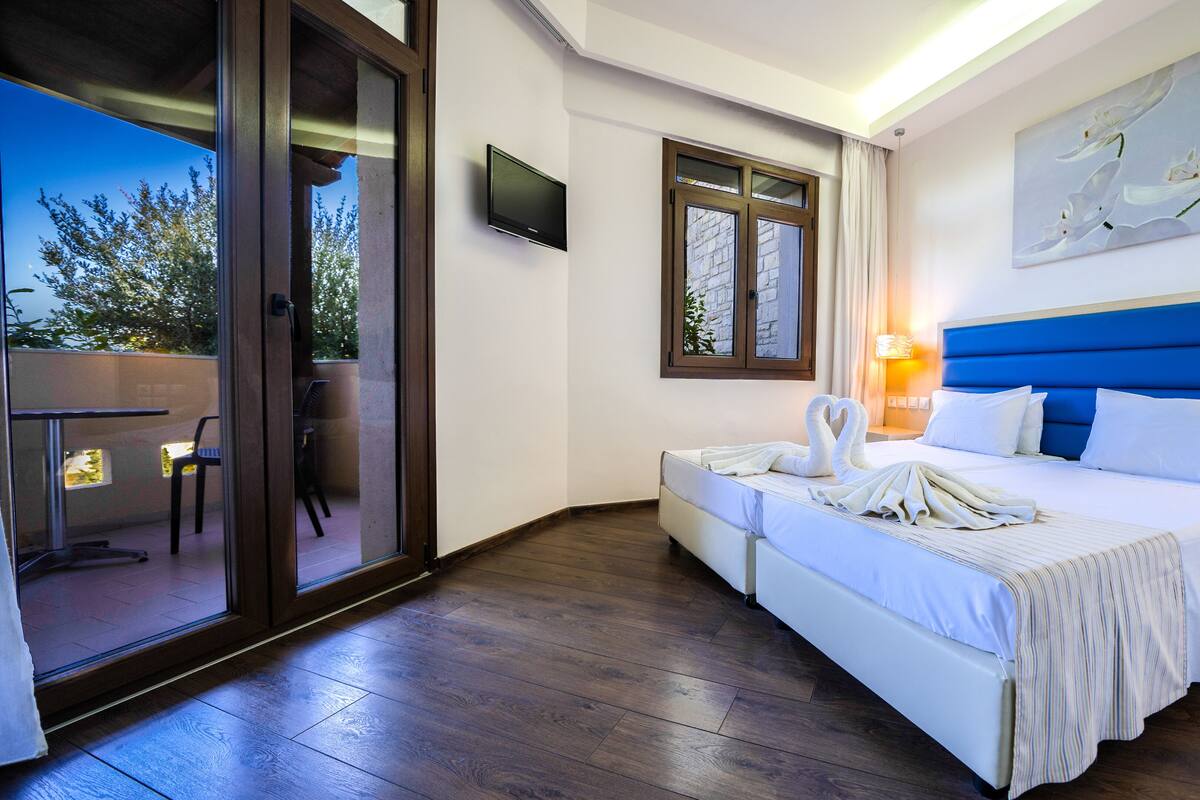 A hotel room with a bed, a TV, a painting, and a view of the outdoors.
