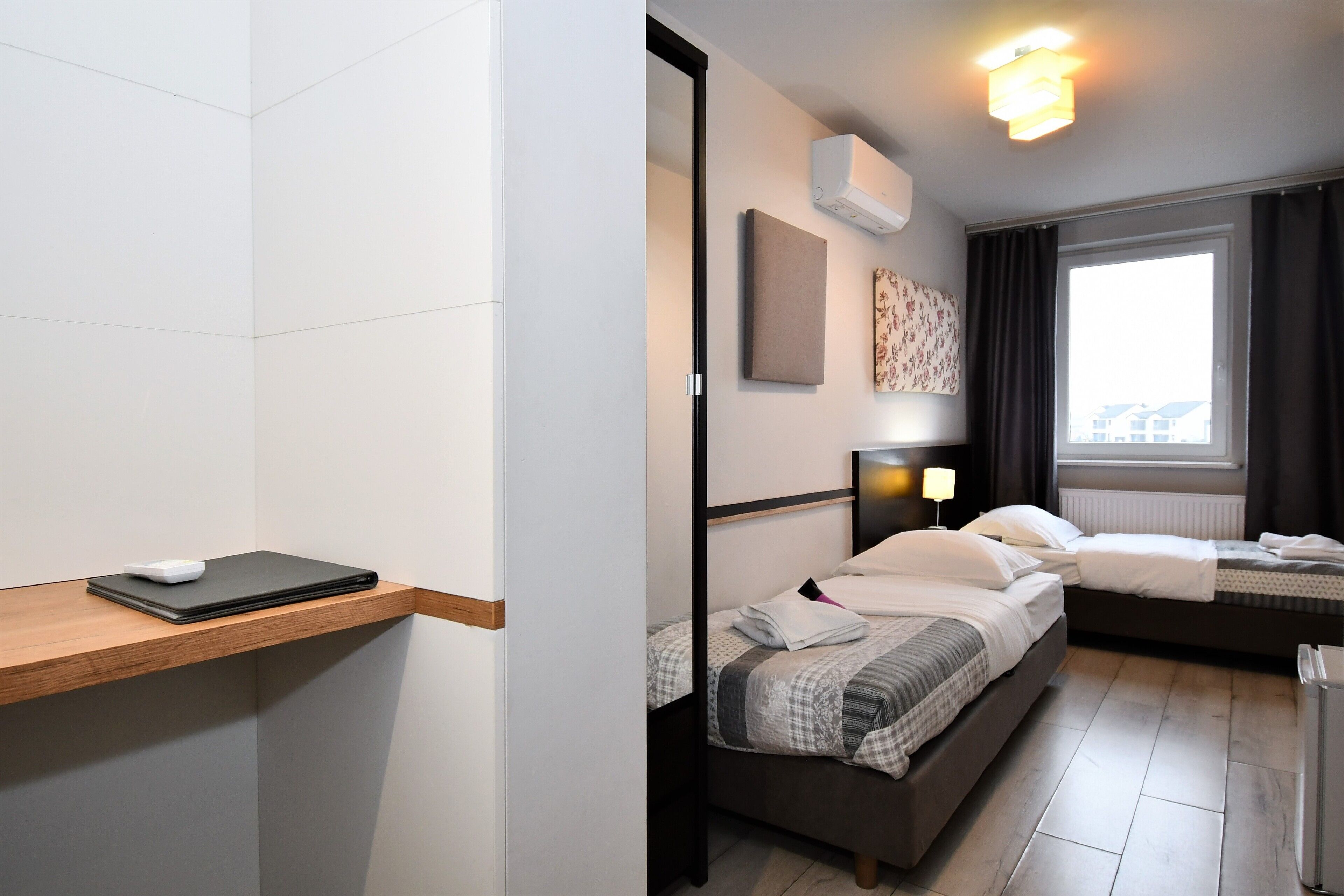 A modern hotel room with a wooden desk, a bed with a headboard, a bedside table, a lamp, and a window with curtains.